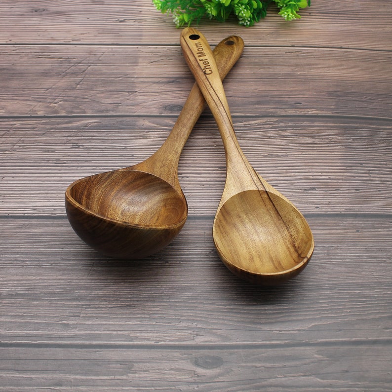 Op de afbeelding: Twee houten soeplepels met een bruine houten afwerking. De lepels zijn over elkaar gekruist. De steel van een lepel heeft de tekst "Chef Mom" gegraveerd.
