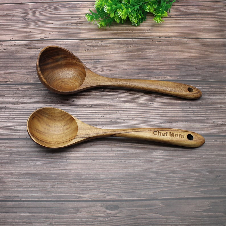 Op de afbeelding: Twee houten lepels, een met een ronde kom en de andere met een ondiepere kom. De lepel met de ondiepe kom heeft de woorden "Chef Mom" gegraveerd op het handvat.