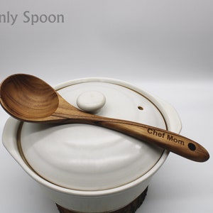 Op de afbeelding: Een houten lepel met een ronde kom en een lange steel. De steel is gegraveerd met de woorden "Chef Mom". De lepel ligt op de deksel van een witte keramische pot.