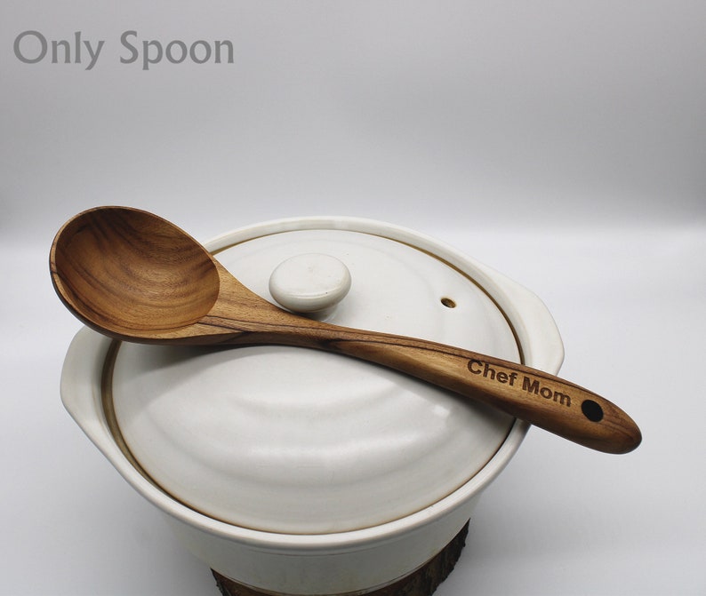 Op de afbeelding: Een houten lepel met een ronde kom en een lange steel. De steel is gegraveerd met de woorden "Chef Mom". De lepel ligt op de deksel van een witte keramische pot.