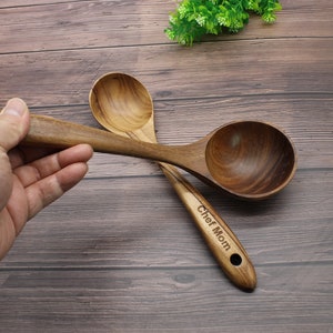 Op de afbeelding: Twee houten serveerscheppen met een bruine houtnerf. De grotere schep heeft een diepe kom en de kleinere schep heeft een ondiepere kom. De grotere schep heeft de tekst "Chef Mom" gegraveerd op het handvat.