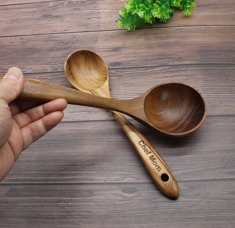 Op de afbeelding: Twee houten serveerscheppen met een bruine houtnerf. De grotere schep heeft een diepe kom en de kleinere schep heeft een ondiepere kom. De grotere schep heeft de tekst "Chef Mom" gegraveerd op het handvat.
