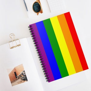 May include: A spiral-bound notebook with a vibrant rainbow flag design. The cover features vertical stripes in the colors of the Pride flag: red, orange, yellow, green, blue, and purple. The notebook is open on a white surface, with a gold paper clip and sunglasses nearby.