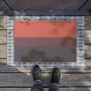 May include: A doormat with a three-color horizontal stripe design. The top stripe is a light orange, the middle stripe is a dark brown, and the bottom stripe is a dark gray. The doormat is on a wooden deck.