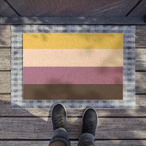 Puede incluir: Un felpudo rectangular con cuatro franjas horizontales en amarillo, beige, malva y marrón oscuro, rodeado por un borde a cuadros grises y blancos. La alfombra está colocada sobre una superficie de madera con un par de zapatillas negras.