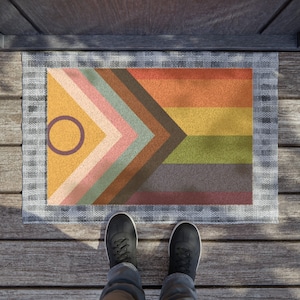 May include: A doormat with a colorful geometric design featuring a rainbow flag with a circle in the upper left corner. The doormat is on a wooden deck.