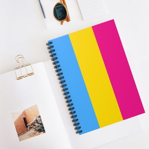 May include: A spiral bound notebook with a blue, yellow, and pink striped cover. The notebook is on a white surface.