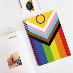Puede incluir: Un cuaderno de espiral con un diseño de bandera del orgullo colorido. La bandera presenta un fondo amarillo con un círculo morado, seguido de rayas blancas, rosas, azul claro, marrones, negras, moradas, azules, verdes, amarillas, naranjas y rojas.