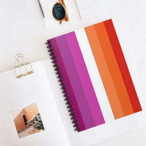 May include: A spiral bound notebook with a lesbian pride flag design. The cover features stripes of pink, purple, white, orange, and red.