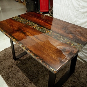 May include: A rectangular wooden table with a glossy finish. The tabletop features a combination of dark wood and a central strip of small stones. The table has black metal legs and sits on a brown rug.