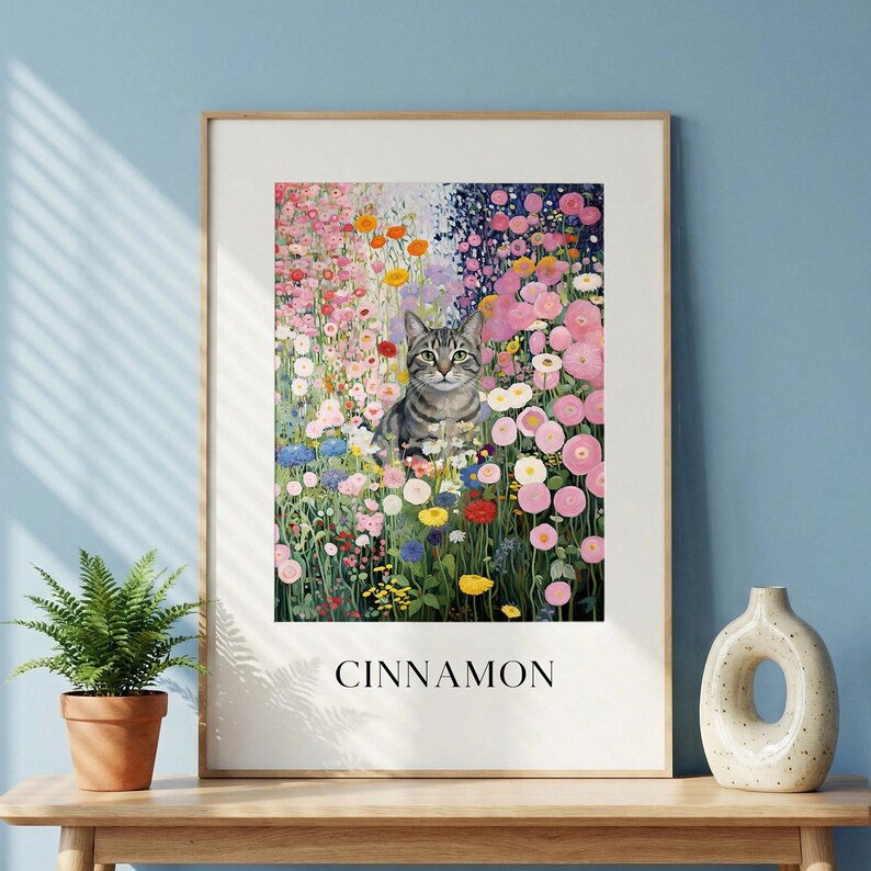 May include: A framed art print featuring a tabby cat amidst a vibrant flower garden. The artwork showcases a variety of colorful blooms in pink, orange, and blue hues. The word "CINNAMON" is printed in a bold font at the bottom of the print.
