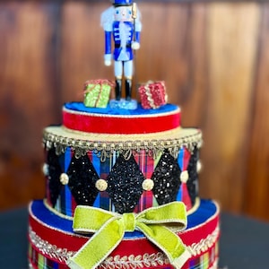 May include: A decorative, tiered Christmas ornament featuring a blue and white nutcracker soldier figurine atop a red, blue, and gold base. The ornament is adorned with plaid patterns, glitter, and a green bow. Two small gift boxes are also present.