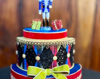 May include: A decorative, tiered Christmas ornament featuring a blue and white nutcracker soldier figurine atop a red, blue, and gold base. The ornament is adorned with plaid patterns, glitter, and a green bow. Two small gift boxes are also present.