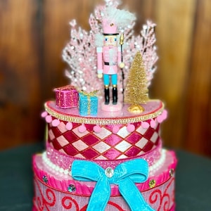 May include: A decorative, tiered Christmas display featuring a pink nutcracker soldier with a white plume, standing atop a pink and red cake-like structure. The display includes a gold tree, pink presents, and a turquoise bow.