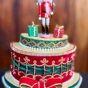 May include: A festive, tiered Christmas cake with a red and green color scheme. The cake is decorated with a red bow, gold trim, and a nutcracker figurine on top. Two small gift boxes are also on top.