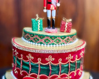 May include: A festive, tiered Christmas cake with a red and green color scheme. The cake is decorated with a red bow, gold trim, and a nutcracker figurine on top. Two small gift boxes are also on top.