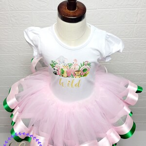 Puede incluir: Un tut&uacute; de tul rosa con ribete de cinta de sat&eacute;n verde y rosa. Una camiseta blanca con un gr&aacute;fico de animales y el texto "Two Wild" se lleva sobre el tut&uacute;.