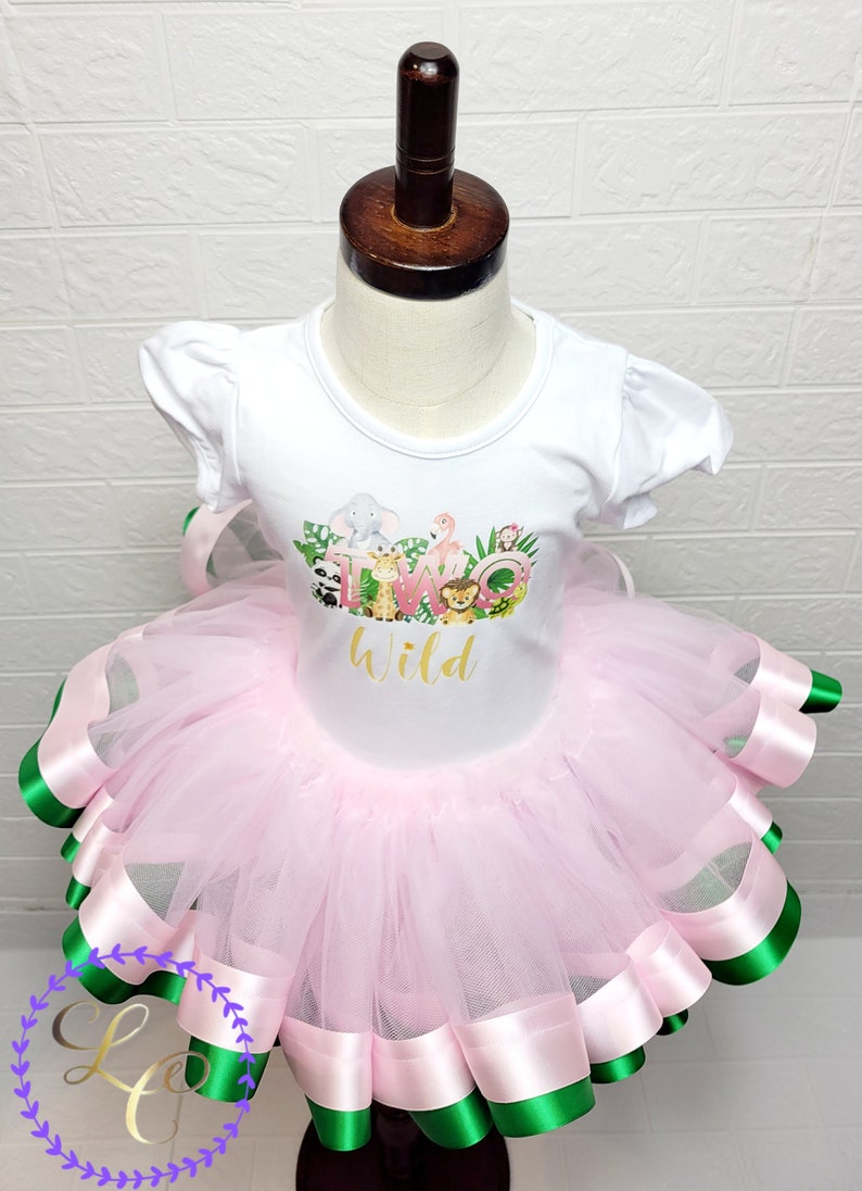Puede incluir: Un tut&uacute; de tul rosa con ribete de cinta de sat&eacute;n verde y rosa. Una camiseta blanca con un gr&aacute;fico de animales y el texto "Two Wild" se lleva sobre el tut&uacute;.