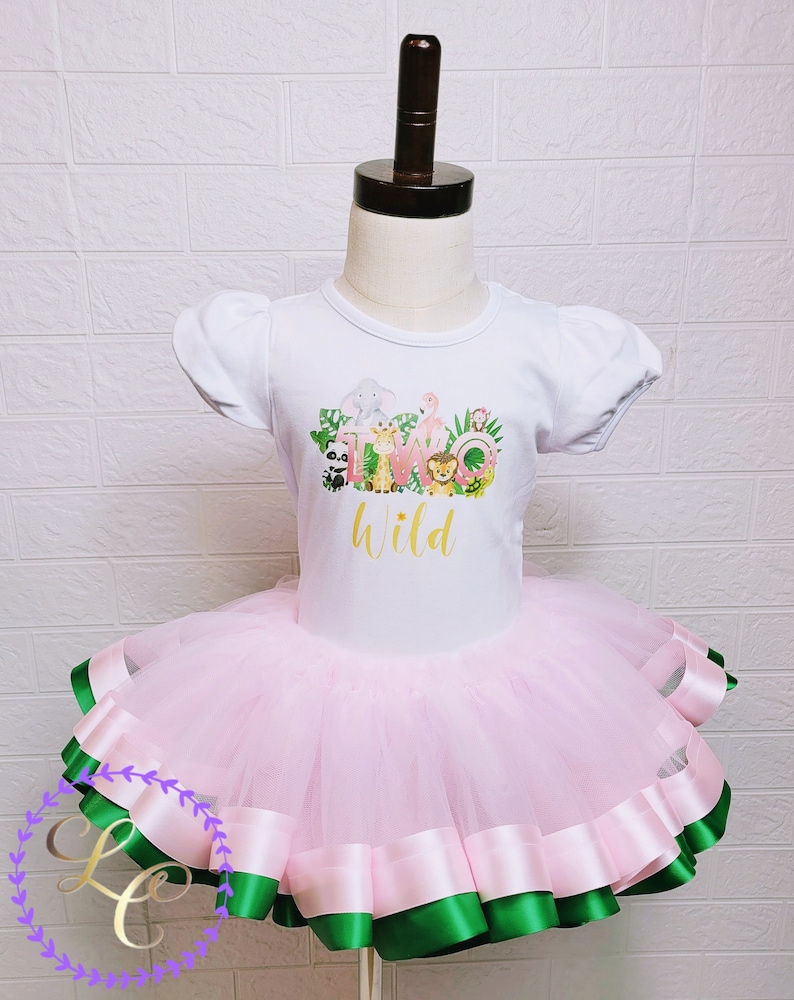 Puede incluir: Un tut&uacute; de tul rosa con ribete de cinta de sat&eacute;n verde y rosa. Una camiseta blanca con un gr&aacute;fico de animales de la selva y el texto "Two Wild" se lleva sobre el tut&uacute;.
