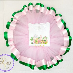 Puede incluir: Falda de tut&uacute; de tul rosa con ribete de cinta verde y rosa. La falda se combina con una camiseta blanca con un gr&aacute;fico de animales de la selva y el texto "DOS".