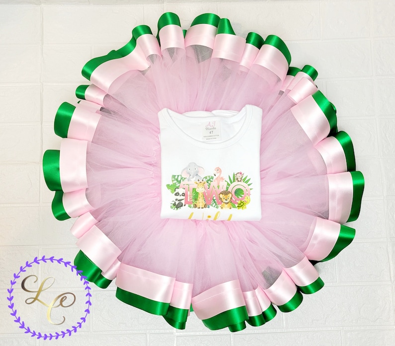 Puede incluir: Falda de tut&uacute; de tul rosa con ribete de cinta verde y rosa. La falda se combina con una camiseta blanca con un gr&aacute;fico de animales de la selva y el texto "DOS".