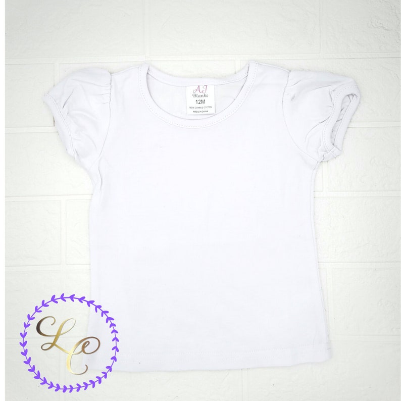 Puede incluir: Camiseta blanca de manga corta con mangas abullonadas. La camiseta tiene una etiqueta que dice "A7 Blanks 12M".
