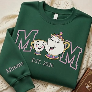 May include: Green sweatshirt with the word "MOM" in pink block letters, featuring a cartoon teacup and teapot. The text "EST. 2026" is below the characters. The sleeve reads "Mimmy" in pink.