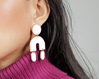 May include: A pair of dangle earrings featuring a small circular stud connected to a larger arch-shaped piece. Two small oval-shaped pendants hang from the bottom of the arch. The earrings are a solid, light cream color with a smooth finish.