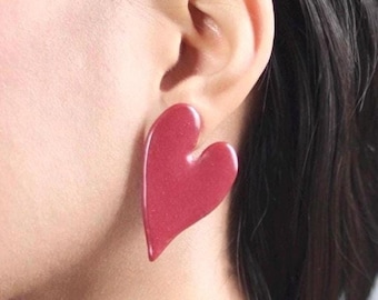 May include: A large, asymmetrical heart-shaped earring with a smooth, glossy finish in a deep, muted red color. The earring features a slightly elongated, curved design that tapers to a point at the bottom, showcasing a subtle, fine-grained texture.