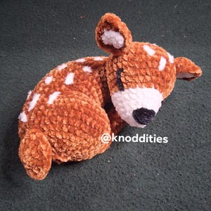 May include: A soft, brown plush deer toy with white spots and a white muzzle. The handmade deer is lying down, with its eyes closed. The deer has a black nose and is made of a textured material.
