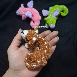 May include: Handmade brown and white spotted crochet creature held in a hand, with two other crochet creatures in the background. One is pink with purple spots and the other is green with purple spots. The text "@knoddities" is visible.