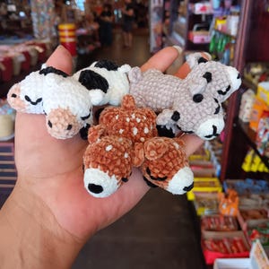 May include: A hand holds several small, crocheted animal figures. The figures include cows in white and black, a gray sheep, and two brown deer with white spots. The animals have black eyes and noses, and white muzzles.