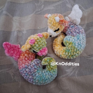 Puede incluir: Dos figuras de dragón de ganchillo hechas de hilo suave y jaspeado en tonos pastel de rosa, amarillo, azul y verde. Cada criatura presenta alas pequeñas, orejas puntiagudas y ojos cerrados. El texto @KnOddities es visible en color blanco.