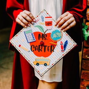 Puede incluir: Un gorro de graduación decorado con una manzana roja, un autobús escolar, una taza de café, una calculadora, lápices de colores y el texto "MS. CARTER".