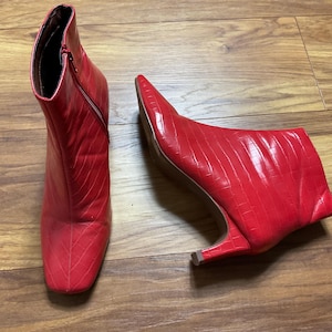 May include: A pair of red ankle boots with a textured, crocodile-like pattern. The boots have a square toe, a side zipper, and a small, curved heel. The boots are displayed on a wooden surface.