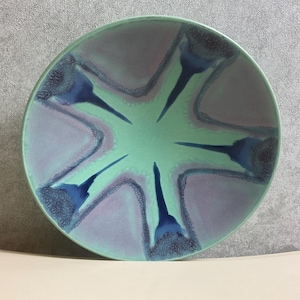 Può includere: Una ciotola rotonda in ceramica con un design unico. La ciotola presenta un centro verde chiaro con un motivo a stella in tonalità blu e viola. Il bordo è verde più scuro.