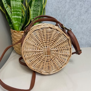 May include: A round, woven rattan handbag with a brown leather strap and handle. The bag has a circular design with a woven pattern and a brown leather accent in the center. The bag is sitting on a white surface.