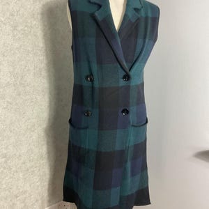 May include: A sleeveless, double-breasted vest in a green, black, and blue plaid pattern. The vest has a notched collar, two front pockets, and black buttons. The garment is displayed on a mannequin.