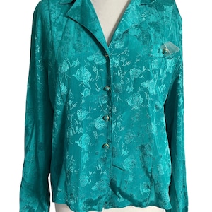 May include: Teal button-down blouse with a floral design. The long-sleeved shirt features a collared neckline and a single pocket. The buttons are gold-coloured. The fabric has a subtle sheen.