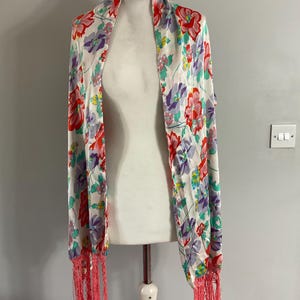 May include: A white silk scarf with a floral print in shades of pink, red, purple, green, and yellow. The scarf has pink fringe on the ends.
