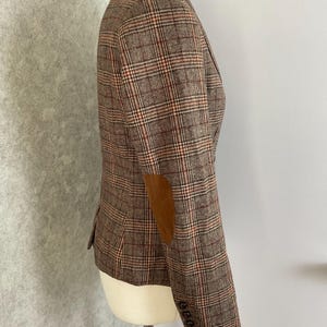 May include: A brown and grey plaid blazer featuring a notched lapel, a single front button closure, two flap pockets, and brown elbow patches. The sleeves have four small buttons at the cuffs, and the interior is lined with a dark patterned fabric.