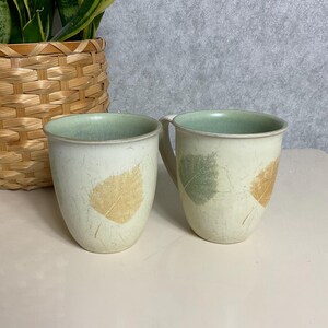 May include: Two off-white ceramic mugs with leaf imprints in gold and green. The mugs have a light green interior and a handle on one. A woven basket with a plant is in the background.