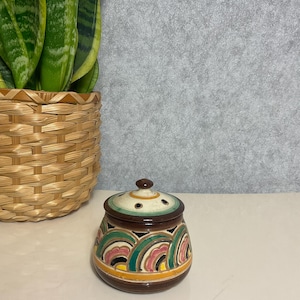 May include: A small, decorative ceramic jar with a lid. The jar is brown with colourful, curved patterns in green, pink, and yellow. The lid is white with brown accents and small holes. A woven basket with a green plant is in the background.