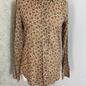 May include: A long-sleeved shirt with a floral print. The shirt has a cream-coloured base covered in small red flowers and green leaves. It features a classic collar and buttoned cuffs. The shirt is buttoned down the front.