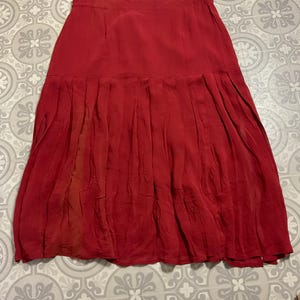 May include: A red skirt with a pleated design. The skirt has a high waist and a flowing, knee-length hem. The fabric appears to be lightweight and has a slight sheen. The skirt is laying flat on a patterned surface.