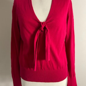 May include: A bright pink sweater with a V-neck and long sleeves. The sweater features a tie detail at the neckline and a ribbed hem. The garment is displayed on a mannequin.