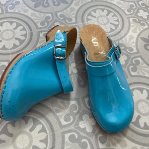 May include: Pair of bright turquoise clogs with a glossy finish. The clogs have a wooden sole with a leather strap and buckle closure. The interior is lined with a light-colored material. The clogs are size 5.