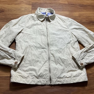 May include: A light beige jacket with a full zipper closure, two front pockets, and buttoned cuffs. The jacket has a collared neckline and a light blue lining visible at the collar. The jacket is made of a lightweight material.