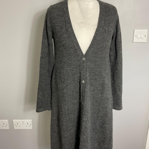 May include: A long, grey cardigan with a V-neck and long sleeves. The sweater has a button closure down the front and is displayed on a mannequin. The fabric appears to be a soft knit.