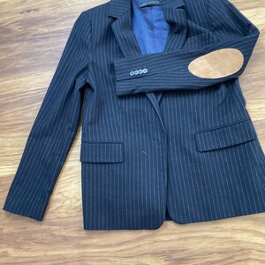 May include: A navy blue pinstriped blazer featuring a notched lapel, two front flap pockets, and a blue interior lining. The sleeve displays four small buttons at the cuff and a tan suede elbow patch. The label inside reads Zara Woman.